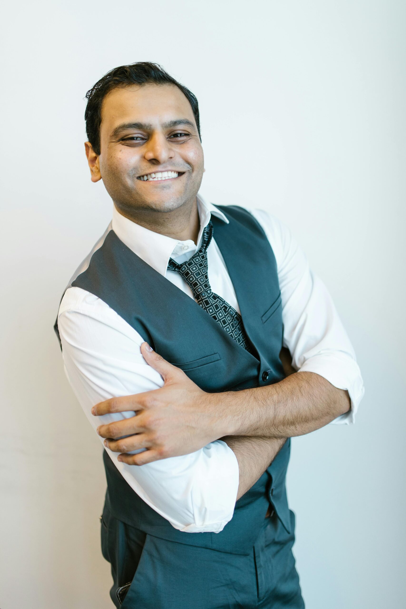 Smiling man in a formal vest and tie, arms crossed, exuding confidence.